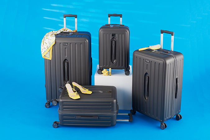 Four charcoal Skyflite Modern Trunk suitcases of varying sizes with spinner wheels and telescopic handles, styled with travel accessories against a bright blue background.