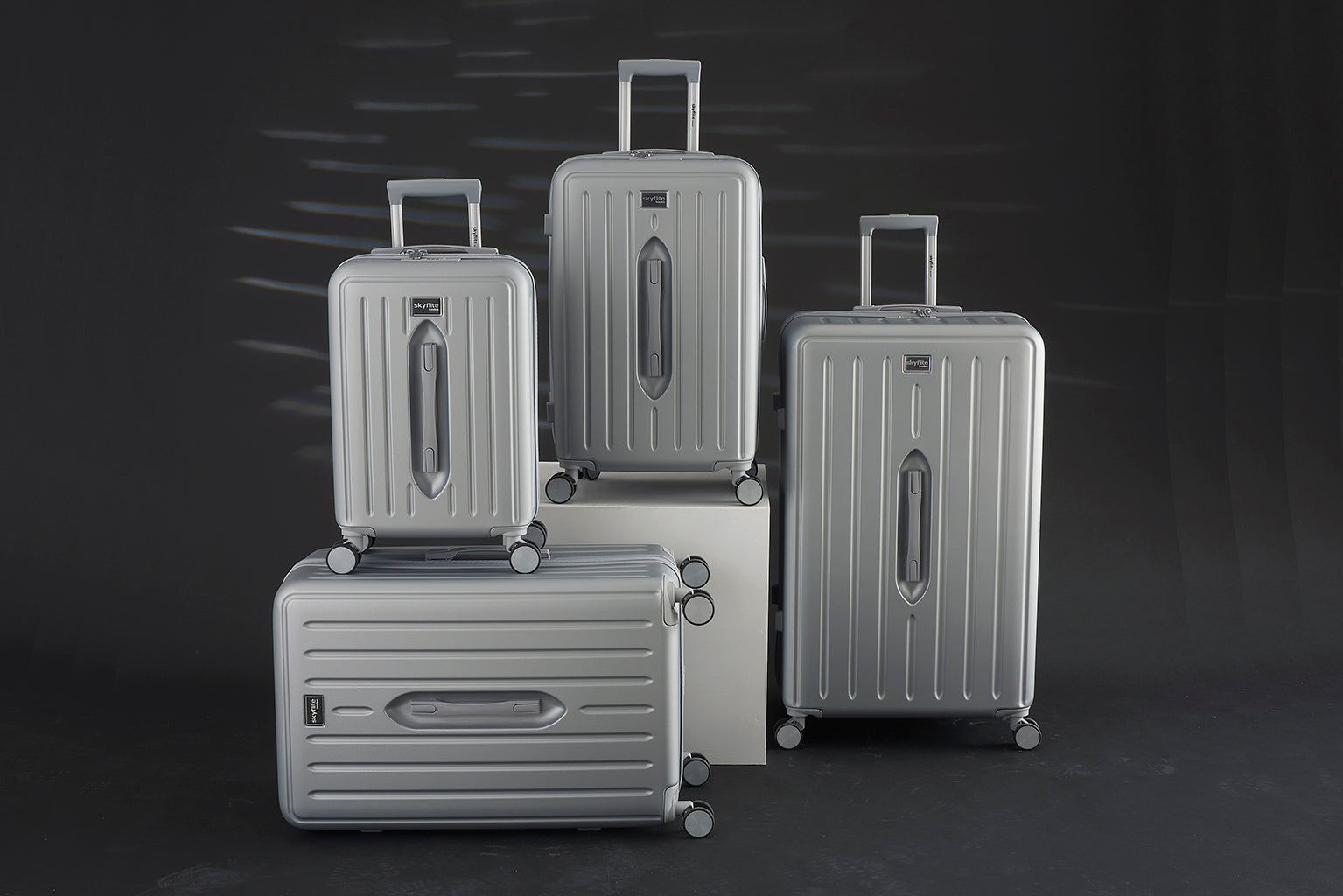 Four silver hard shell suitcases of different sizes with spinner wheels and telescopic handles, displayed against a dark background. The luggage features a modern, sleek design with vertical ridges and Skyflite branding.